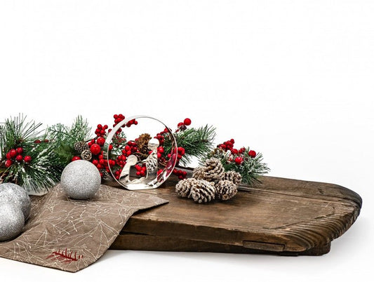 presépio Nazaré decorativo em vidro junto a pinhas, bolas prateadas e raminhos natalícios sobre tábua rústica