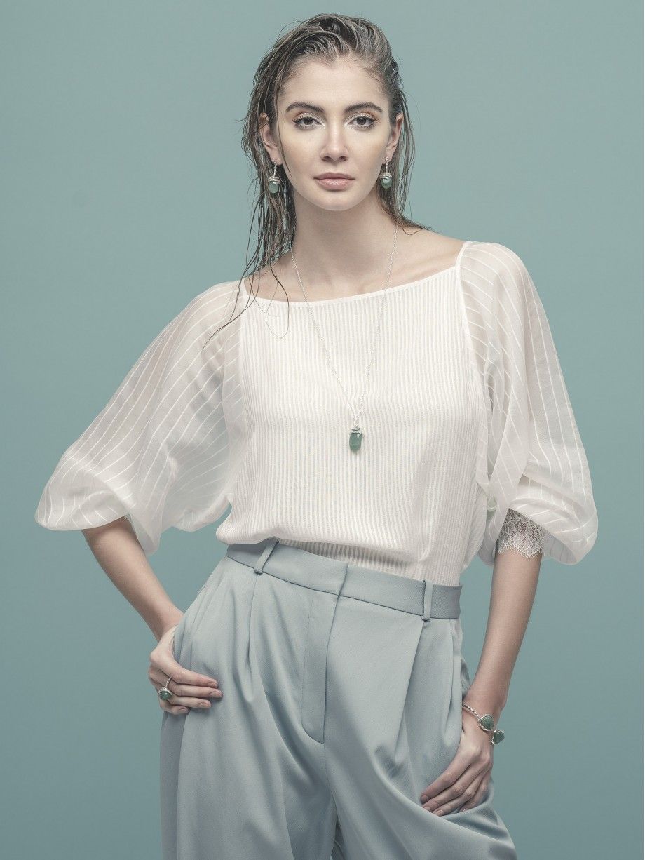 Mulher com brincos naturais femininos verdes e colar Carmo Joalheiros usando blusa branca e calcas azul